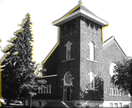 Picture of the Corunna United Methodist Church. A red brick building with white trim. Two round stained-glass windows feature prominently.