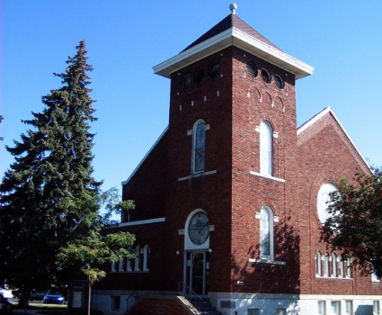 Picture of the Corunna United Methodist Church in color. Red brick with white trim. The picture shows two prominent round stained-glass windows.